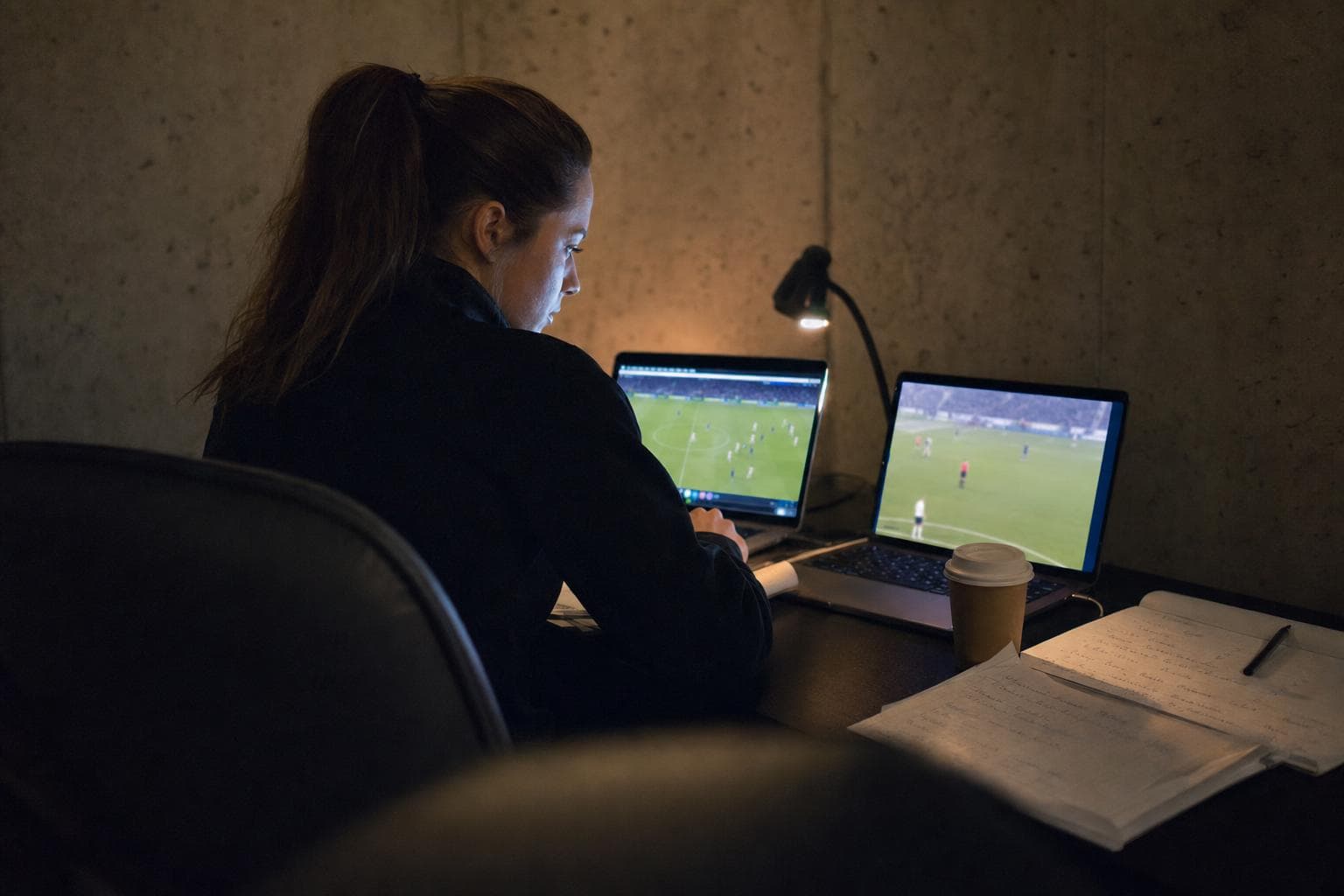 A performance analyst reviewing match footage on two laptops in a dim video room before dawn